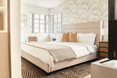 Modern bedroom with beige upholstered headboard, palm pattern wallpaper, nightstand, mirror, armchair, pillows, blankets, nightstand.